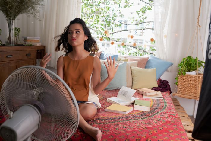 A young woman sits on her bed in front of a fan, fanning herself with her hands.